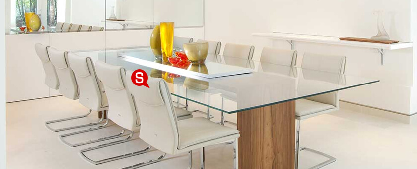 Jadalnia w stylu minimalistycznym ze szklanym, transparentnym stołem na drewnianej podstawie oraz białymi krzesłami. Całości aranżacji dopełniają dekoracje w ciepłych kolorach.